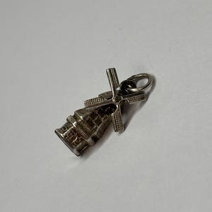 May include: A small, silver-colored windmill charm with detailed brickwork on the base and a cross-shaped rotor. The charm has a loop for attaching to a necklace or bracelet. The windmill's blades are positioned to catch the wind.