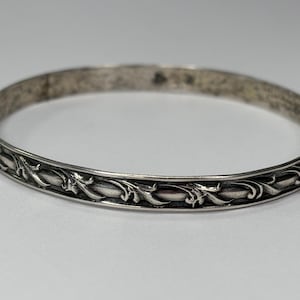 Puede incluir: Un brazalete plateado con un diseño floral ornamentado y repetitivo. El brazalete es circular y parece estar hecho de metal. El patrón floral está en relieve y detallado, agregando textura a la pieza. El brazalete se muestra sobre un fondo neutro.
