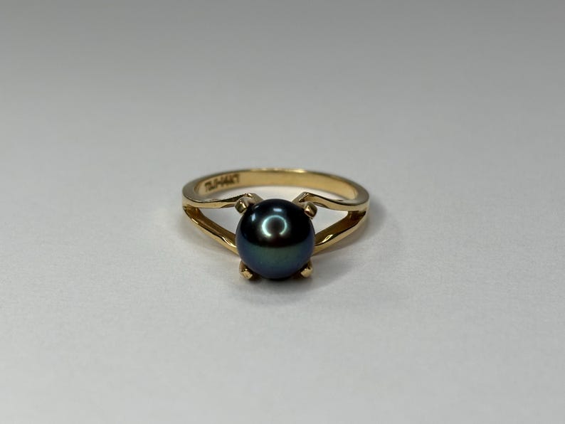 Puede incluir: Un anillo de oro con una perla oscura e iridiscente. El anillo tiene un dise&ntilde;o de ca&ntilde;a dividida, con la perla sujeta por cuatro garras. El anillo est&aacute; sobre un fondo neutro.