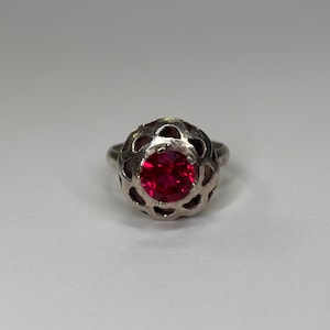May include: A silver ring with a bright red gemstone at the center. The ring's setting has an open, floral-like design around the gemstone. The ring is set against a plain white background, highlighting its intricate details and vibrant color.