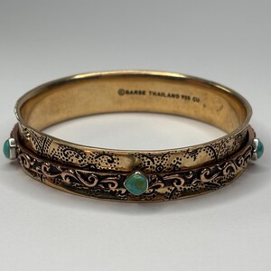 Puede incluir: Un brazalete de oro con una banda de cobre y piedras turquesa. El brazalete está grabado con un diseño floral y las palabras "Saras Thailand 925 Cu".