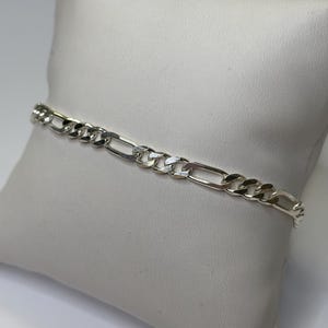 Könnte beinhalten: Ein silberfarbenes Figaro-Kettenarmband. Das Armband hat ein Muster aus drei kleinen kreisförmigen Gliedern, gefolgt von einem länglichen ovalen Glied. Das Armband wird auf einer weißen Oberfläche präsentiert.