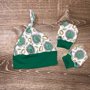 May include: A baby hat and two pairs of mittens. The hat has a green brim and a white top patterned with green hobbit door designs. The mittens have green cuffs and match the hat's pattern. The set is designed for infants.