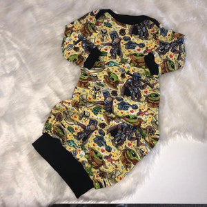May include: A yellow long-sleeved baby onesie with a black trim. The onesie features a repeating pattern of cartoon characters from the Star Wars franchise, including Baby Yoda, Mandalorian, and other characters.