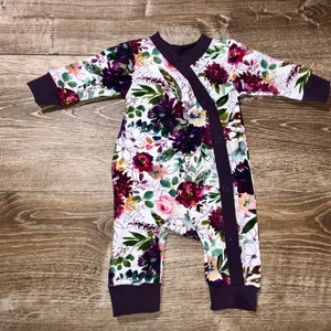 May include: A white long-sleeved baby onesie with a floral print in shades of purple, pink, and green. The onesie has a purple trim and snap closure.