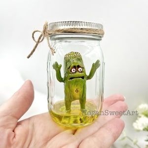 May include: A clear glass jar with a silver metal lid and a tied twine bow containing a small, green, anthropomorphic pickle figure with wide eyes and raised arms, standing in a yellow liquid, with the text HoganSweetArt visible.
