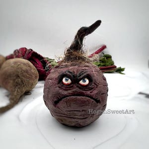 May include: A hand-crafted beet with a grumpy face, painted with red, blue and brown details. The beet has a long, dark brown stem and is surrounded by green beet leaves.
