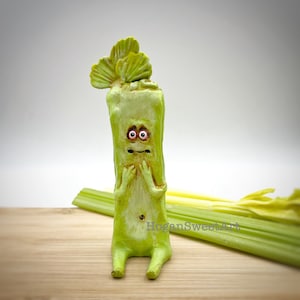 May include: A green celery figurine with a surprised expression, sitting on a wooden surface with celery stalks in the background. The figurine has large eyes and a small mouth.