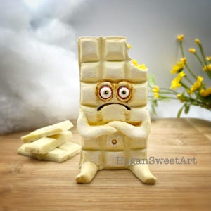 May include: A quirky, off-white chocolate bar figure with a downcast expression, arms folded, and legs. The bar has a bite taken out, showing a yellow interior. Several chocolate pieces are nearby. The artist's name, "HoganSweetArt", is visible.