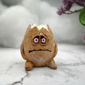 May include: A quirky ceramic egg sculpture, hand-painted with a distressed finish. The egg has a cracked top, showing a yellow yolk. It has a sad face with red-rimmed eyes and a downturned mouth. A unique art piece, it is a decorative item.