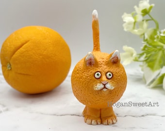 May include: A small orange cat figurine with white fur details. The cat is sitting on a white surface with a whole orange in the background.
