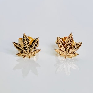 Puede incluir: Un par de pendientes de color dorado con un diseño detallado de hoja de marihuana.