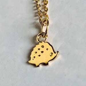 Könnte beinhalten: Eine goldene Halskette mit einem kleinen Anhänger in Form eines Igels. Der Anhänger ist mit einer strukturierten Oberfläche und einem kleinen, runden Auge verziert.