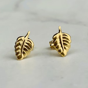 Puede incluir: Un par de pendientes de botón de hoja de oro. Los pendientes son pequeños y delicados, y presentan un diseño simple de hoja.