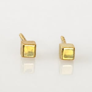 May include: A pair of gold-toned stud earrings, each featuring a small, polished cube-shaped design. The earrings are oriented with the post extending outward, showcasing the smooth, reflective surfaces of the metallic square faces.