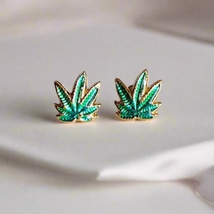 Puede incluir: Un par de pendientes de tono dorado con diseños de hojas de marihuana en esmalte verde.