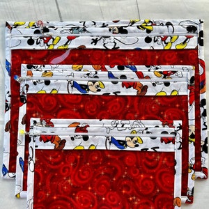 Puede incluir: Conjunto de cuatro bolsas transparentes con cremallera con tejido rojo y blanco. El tejido presenta un patr&oacute;n repetido de personajes de Mickey Mouse. Las bolsas est&aacute;n anidadas una dentro de la otra, con la bolsa m&aacute;s grande en la parte inferior y la m&aacute;s peque&ntilde;a en la parte superior.