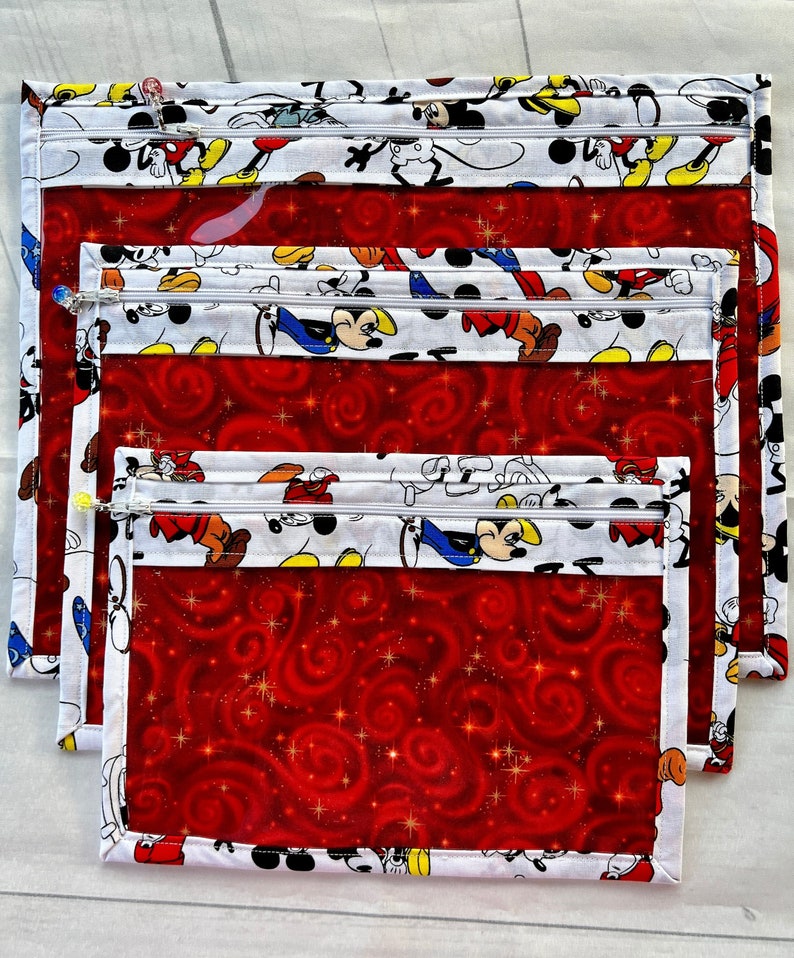 Puede incluir: Conjunto de cuatro bolsas transparentes con cremallera con tejido rojo y blanco. El tejido presenta un patr&oacute;n repetido de personajes de Mickey Mouse. Las bolsas est&aacute;n anidadas una dentro de la otra, con la bolsa m&aacute;s grande en la parte inferior y la m&aacute;s peque&ntilde;a en la parte superior.