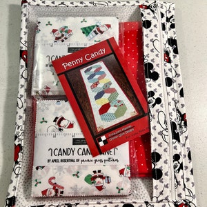 Puede incluir: Una bolsa blanca con cremallera con un patr&oacute;n de Mickey Mouse. La bolsa est&aacute; abierta y contiene una tela roja, una bolsa de pl&aacute;stico transparente con cuadrados de tela y un patr&oacute;n impreso para una colcha Penny Candy. El patr&oacute;n se titula "Penny Candy" y es de Tricia Lynn Maloney, Orphan Quilt Designs. Los cuadrados de tela son para una colcha Candy Cane de April Rosenthal de Prairie Grass Patterns.