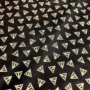 May include: A black fabric featuring a repeating pattern of light yellow Deathly Hallows symbols. Each symbol consists of a triangle containing a vertical line and a circle, arranged in a consistent grid across the entire surface of the material.