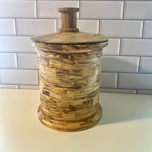 May include: A cylindrical wooden canister featuring a mosaic pattern of small, light and dark brown wood blocks. The container has a matching circular lid with a central knob handle and a flared base, showcasing natural wood grain variations.