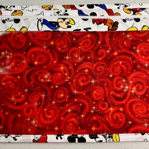 Puede incluir: Una bolsa blanca con cremallera y una ventana de pl&aacute;stico transparente. La bolsa est&aacute; decorada con un patr&oacute;n de remolinos rojo y dorado e im&aacute;genes de Mickey Mouse.