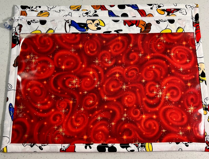 Puede incluir: Una bolsa blanca con cremallera y una ventana de pl&aacute;stico transparente. La bolsa est&aacute; decorada con un patr&oacute;n de remolinos rojo y dorado e im&aacute;genes de Mickey Mouse.