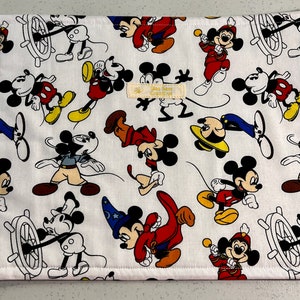 Puede incluir: Salvamanteles de tela blanca con un patr&oacute;n repetido de Mickey Mouse en varias poses. Mickey lleva pantalones cortos rojos, zapatos amarillos y guantes blancos. Algunos personajes de Mickey Mouse llevan sombreros, incluyendo un sombrero de marinero, un sombrero de mago y un sombrero de director de orquesta. El salvamanteles tiene un fondo blanco con una textura sutil.