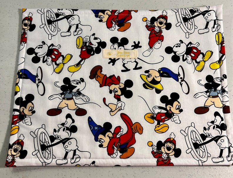 Puede incluir: Salvamanteles de tela blanca con un patr&oacute;n repetido de Mickey Mouse en varias poses. Mickey lleva pantalones cortos rojos, zapatos amarillos y guantes blancos. Algunos personajes de Mickey Mouse llevan sombreros, incluyendo un sombrero de marinero, un sombrero de mago y un sombrero de director de orquesta. El salvamanteles tiene un fondo blanco con una textura sutil.