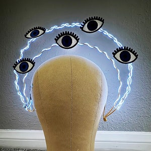 May include: A gold wire headband with five blue evil eye charms. The headband is lit with white LED lights.