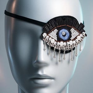May include: An eye patch with a black strap and a decorative eye design. The eye features a blue iris, white and brown beaded details, and a black border. Dangling silver chains with small clear beads hang from the bottom.