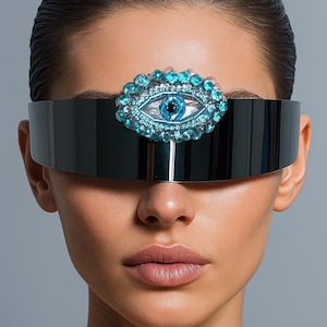 May include: Black visor-style sunglasses with a decorative eye-shaped embellishment. The embellishment features a blue eye design surrounded by blue gemstones. The sunglasses are worn by a person with smooth skin and dark hair.
