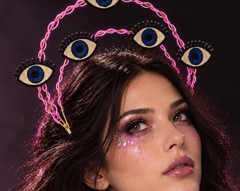 May include: A halo-style headpiece featuring a bright pink twisted wire frame adorned with four eye-shaped ornaments. Each ornament has a black border, a gold-toned iris, and a dark blue pupil, arranged symmetrically along the curved wire structure.