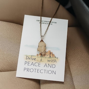 May include: A necklace with a light-colored stone pendant wrapped in wire, suspended from a bronze-colored chain. The pendant hangs over a card with the text "Drive with PEACE AND PROTECTION" and a desert landscape illustration.