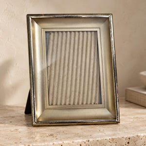 May include: A rectangular picture frame with a metallic gold finish and a distressed silver-toned edge. The frame features a wide, beveled border surrounding a central glass pane that displays a vertical, light brown corrugated paper insert.