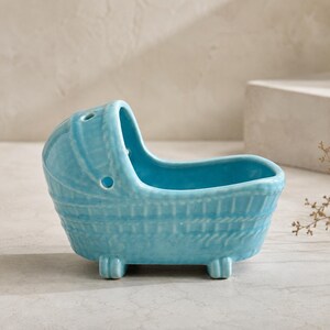 May include: A light blue ceramic planter shaped like a vintage baby bassinet. The vessel features a glossy finish, a textured woven pattern on the exterior, and two small circular openings on the hood section, supported by four short feet.