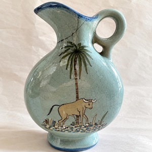May include: A light blue ceramic pitcher with a crackle glaze finish, featuring a painted illustration of a brown bull standing beneath a tall palm tree. The pitcher has a rounded body, a curved handle, and a blue-rimmed spout and base.