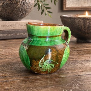 May include: A small, rounded ceramic pitcher featuring a glossy glaze in shades of deep brown and vibrant green. The vessel has a single side handle and a hand-painted, stylized floral motif in green on the front body, with a wide, open rim.