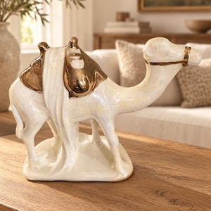 May include: A ceramic camel figurine featuring a glossy, iridescent cream-colored finish. The camel is adorned with metallic gold-toned saddle accents and a matching gold-toned bridle strap around its head, standing on a solid base.