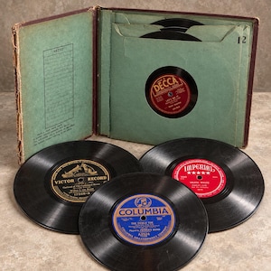 May include: A vintage record album storage book with green paper sleeves containing black vinyl records. Three loose records are displayed in front, featuring circular labels in red, blue, and gold with text including Decca, Columbia, Victor, and Imperial.