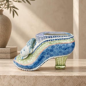 May include: A ceramic shoe-shaped planter featuring a blue and light green color palette with hand-painted swirl and dot patterns. The design includes a decorative bow on the front, a scalloped collar, and a sturdy heel, all finished with fine detailing.