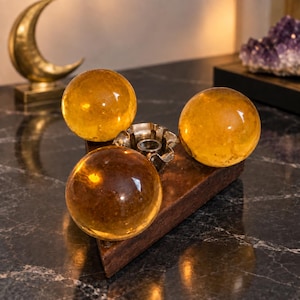 May include: A triangular wooden base holds three smooth, translucent amber-colored spheres arranged around a central silver-toned metal candle holder. The spheres are polished and round, reflecting light across their surfaces, while the wooden base has a dark, textured finish.