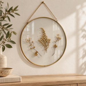 May include: A circular wall hanging featuring a thin brass frame and clear glass panes. Inside the glass, three dried, light-colored botanical stems are pressed and displayed. The frame is suspended by a matching brass chain for wall mounting.