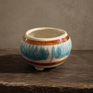 May include: A small, round, off-white ceramic bowl featuring a hand-painted design of horizontal rust-colored bands and a central row of blue, petal-like shapes. The bowl has a smooth, glazed finish and rests on three small, integrated feet.