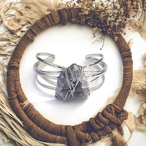 May include: A silver wire wrapped bracelet with a large amethyst crystal in the center. The bracelet is displayed on a brown woven background.