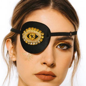 May include: Black eye patch with a gold and crystal embellished eye design. The eye patch is secured with a black elastic band. The eye patch is a fashion accessory.