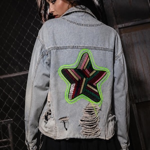 May include: A light blue denim jacket featuring a large, multi-colored star patch on the back. The star is outlined in neon green thread and filled with diagonal stripes of sequins in red, gold, green, and silver. The jacket has distressed, frayed areas.