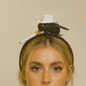 May include: A black fly wearing a white hat and smoking a cigarette, perched atop a black headband. The fly has detailed wings and a small white hat. The cigarette has an orange filter. The headband is worn on a person's head.