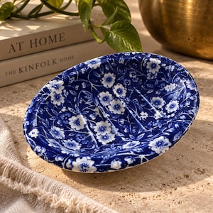May include: An oval soap dish featuring a deep blue and white floral pattern. The dish has a smooth, glossy finish with three raised ridges along the interior base to allow for drainage. The design consists of small, repeating white flowers.