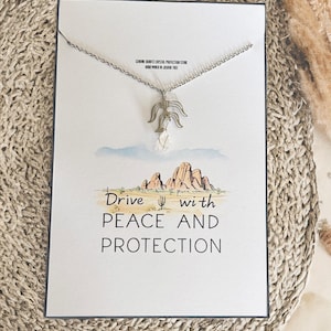May include: A silver necklace with a clear quartz crystal pendant and a silver charm that says "Drive with Peace and Protection". The charm is shaped like a mountain range with a cactus in the foreground. The necklace is displayed on a card with a watercolor background.